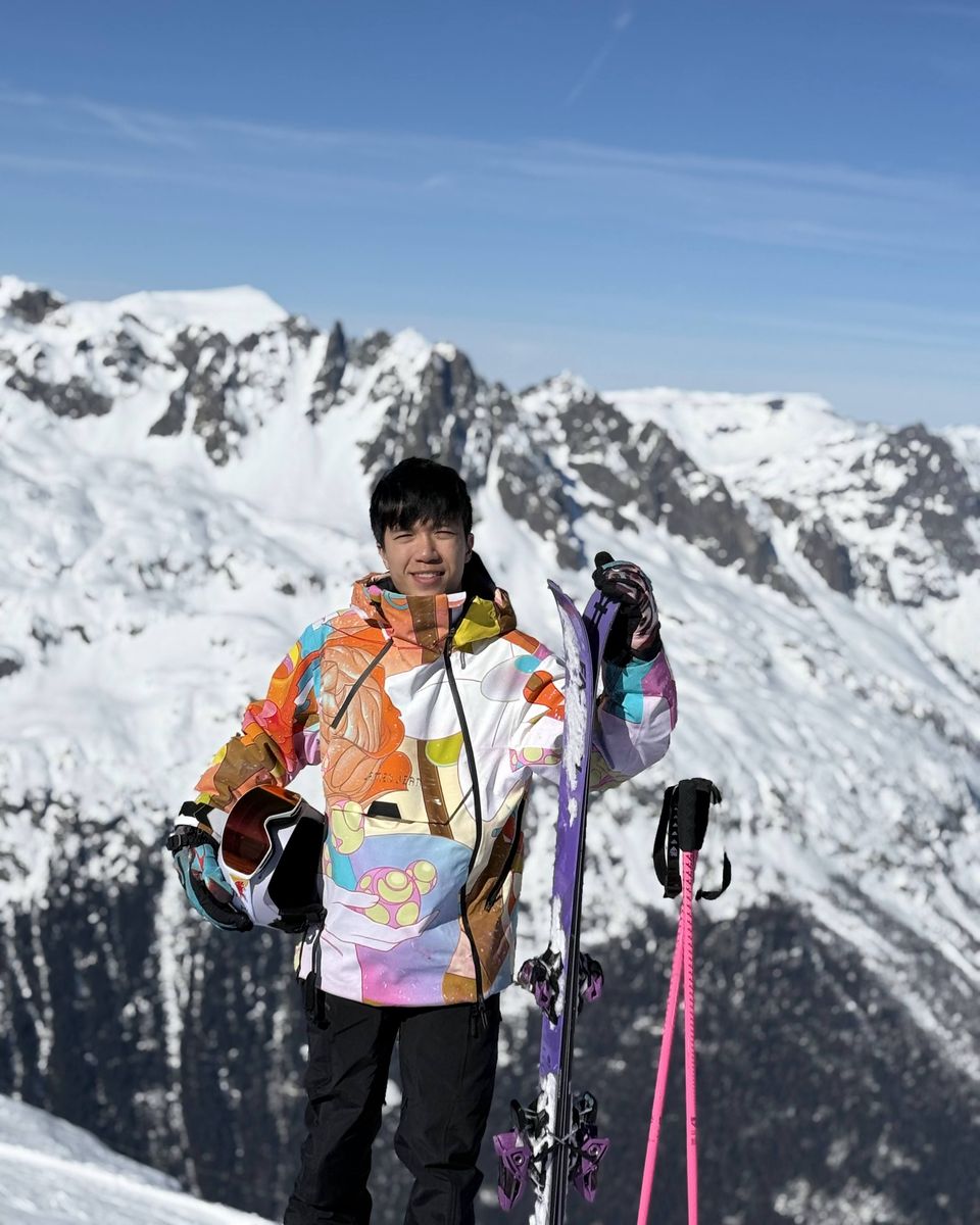 Angus Yip on a snowy summit in a brightly patterned ski jacket, holding a purple ski, pink poles beside him and jagged peaks behind.
